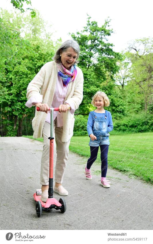 Cheerful senior woman riding push scooter while granddaughter walking in park color image colour image outdoors location shots outdoor shot outdoor shots day