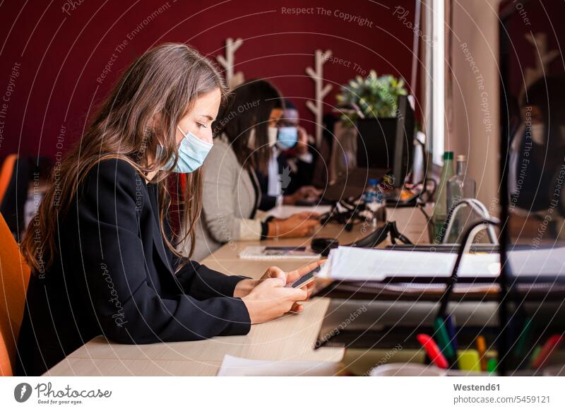 Young businesswoman in protective face mask text messaging through smart phone at desk in office during pandemic color image colour image businesswomen
