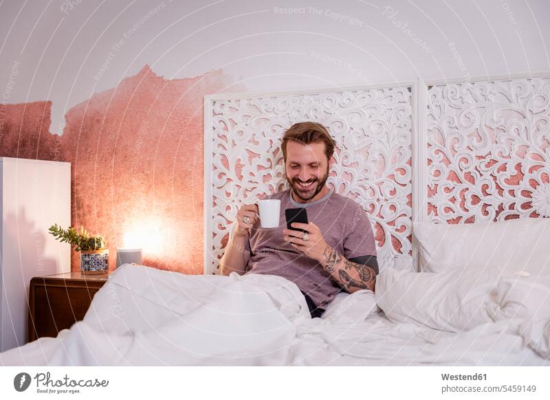 Smiling man holding coffee mug using smart phone while relaxing on bed at home color image colour image leisure activity leisure activities free time