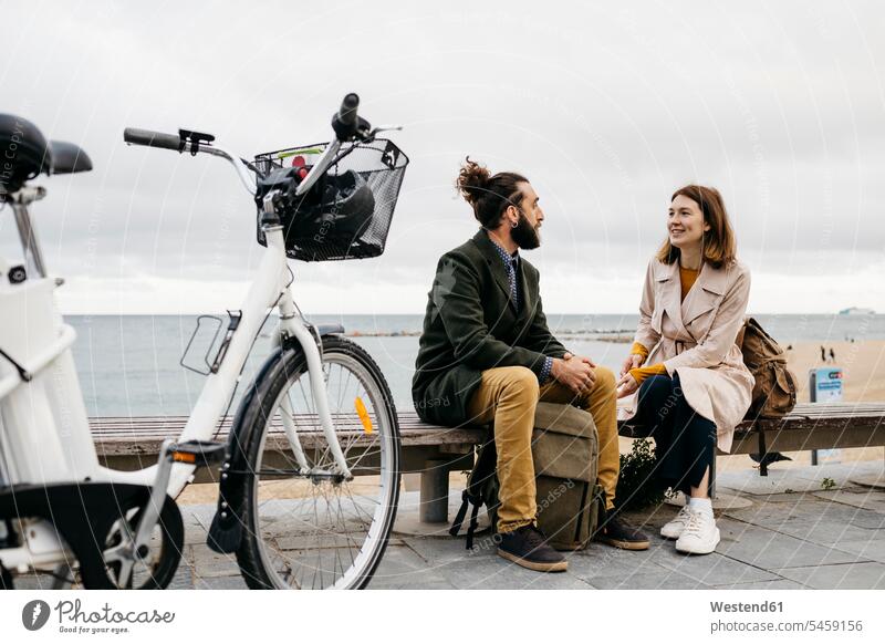 Couple sitting on a bench at beach promenade next to e-bike talking sea front boardwalk city town cities towns E-Bike Electric bicycle Electric Bike speaking