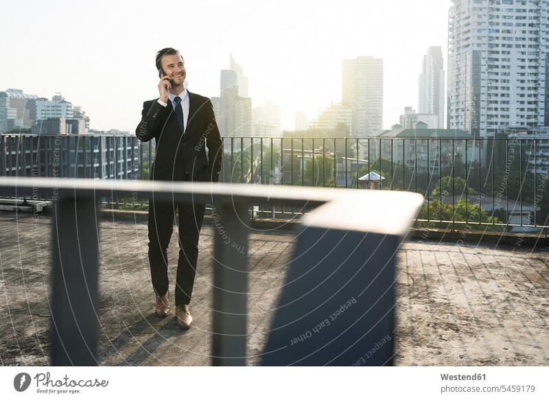 Business man in dark suit speaking into smartphone on city rooftop Fullsuit suits full suit Businessman Businessmen Business men Railing Railings house top