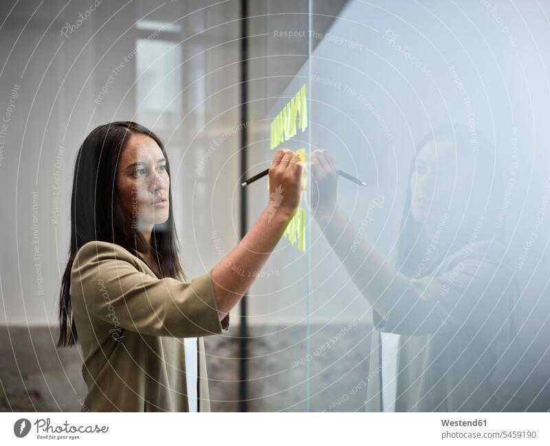 Businesswoman writing on sticky notes at glass pane in office businesswoman businesswomen business woman business women write glass panes offices office room