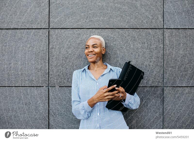 Smiling businesswoman with folder and phone against building in city color image colour image outdoors location shots outdoor shot outdoor shots day