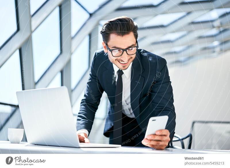 Smiling businessman looking at cell phone at desk in modern office mobile phone mobiles mobile phones Cellphone cell phones desks Businessman Business man