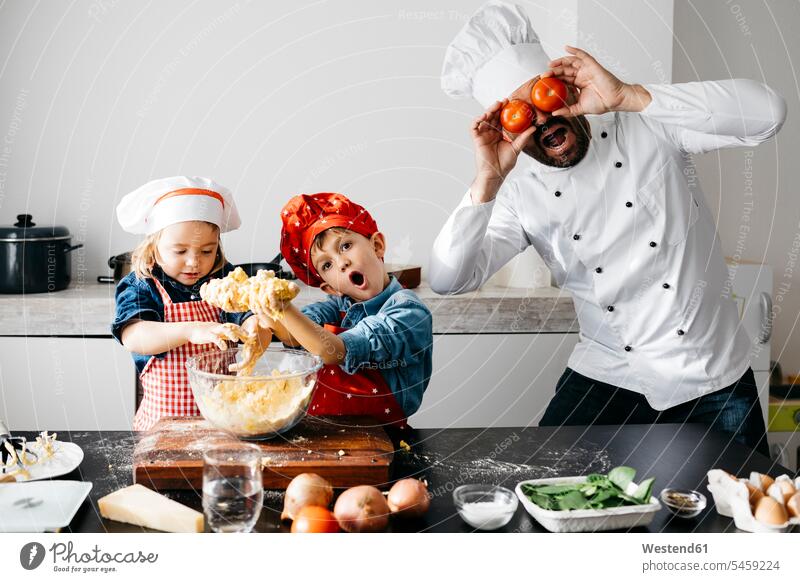 Playful father with two kids covering his eyes with tomatoes in kitchen Chefs cook cooks Bowls hold learn smile delight enjoyment Pleasant pleasure Cheerfulness