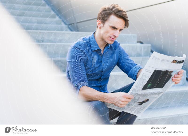 Confident handsome businessman reading newspaper while sitting on steps color image colour image Germany outdoors location shots outdoor shot outdoor shots day