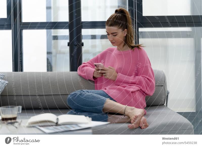 Young woman sitting barefoot on the couch using cell phone couches settee settees sofa sofas telecommunication phones telephone telephones cell phones Cellphone