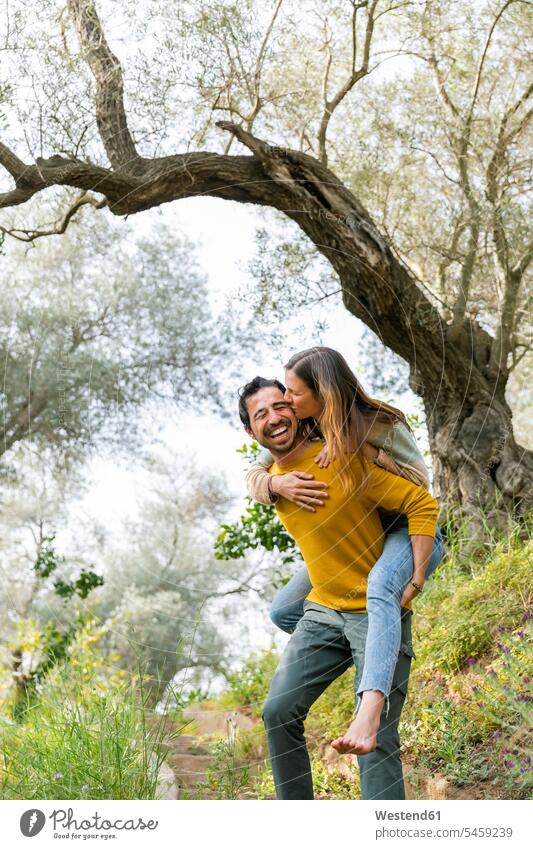 Cheerful man piggybacking romantic girlfriend while standing against trees at countryside color image colour image Spain leisure activity leisure activities