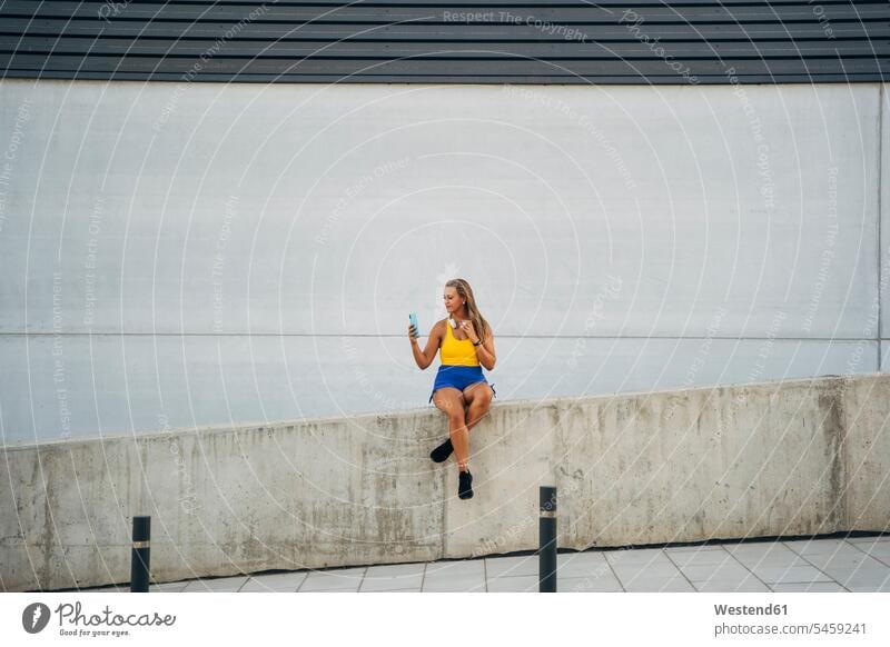 Blond athletic woman taking a selfie, sitting on wall headphone headset telecommunication phones telephone telephones cell phone cell phones Cellphone mobile