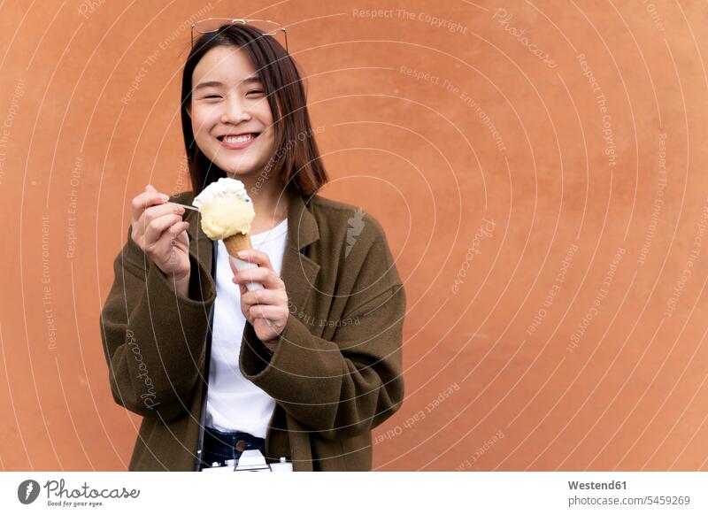 Happy young woman eating an ice cream cone at an orange wall Asian Ethnicity Asians cooling Cool Down cooling down Italy female Asian female Asians