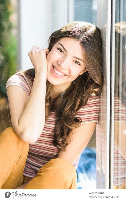 Portrait of smiling young woman sitting at the window at home windows portrait portraits females women smile Seated Adults grown-ups grownups adult people