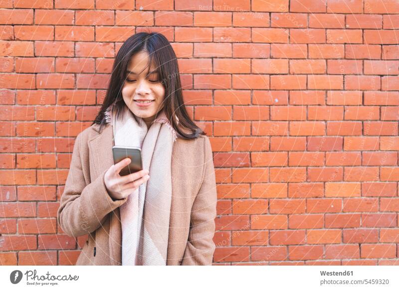 Smiling young woman using cell phone at brick wall mobile phone mobiles mobile phones Cellphone cell phones smiling smile females women brick walls telephones