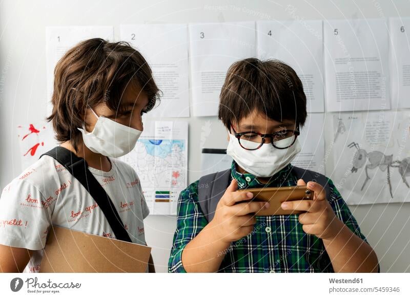 Boy wearing mask looking at friend using smart phone while standing against wall in school color image colour image Spain 10-11 years 10 to 11 years children
