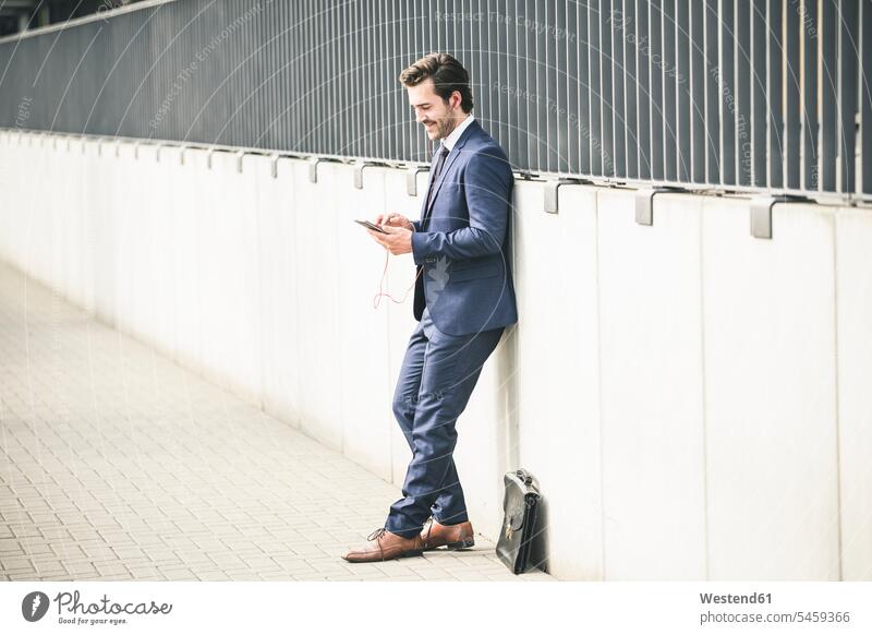 Businessman leaning against a wall in the city checking cell phone freelancer freelancing business business world business life Instant Messaging Messenger