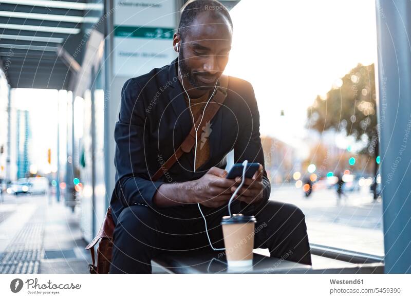 Young businessman sitting at tram stop in the evening using earphones and smartphone human human being human beings humans person persons African black