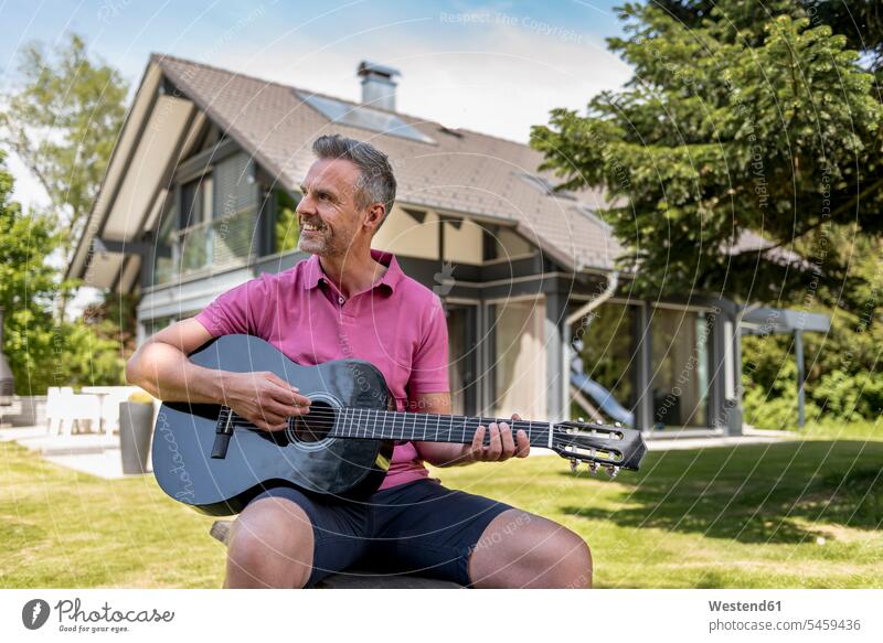 Smiling mature man sitting in garden of his home playing guitar men males guitars house houses smiling smile Seated gardens domestic garden Adults grown-ups