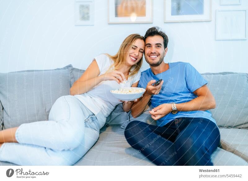 Smiling couple watching TV while enjoying popcorn on sofa at home color image colour image indoors indoor shot indoor shots interior interior view Interiors day