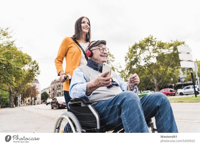 Young woman pushing senior man with headphones and smartphone in wheelchair human human being human beings humans person persons caucasian appearance