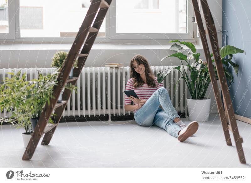 Woman sitting on ground in her new home, reading e-book, surrounded by plants ladder ladders Sitting On The Floor Sitting On Floor woman females women at home