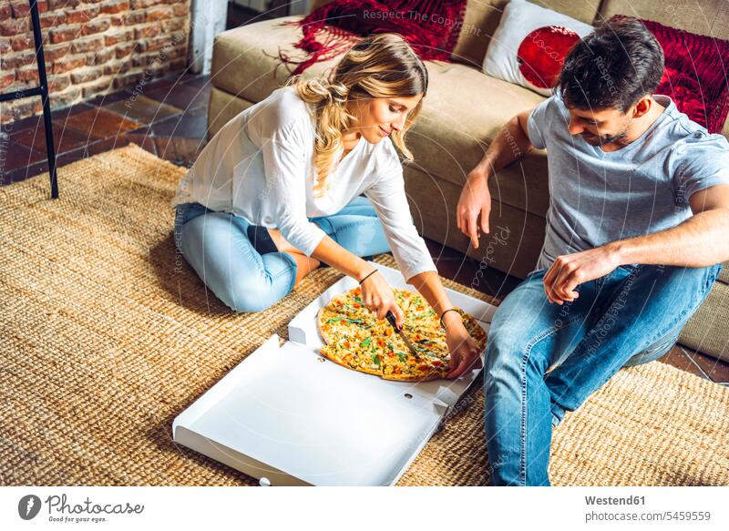 Young couple sitting on floor of living room and cutting pizza in box boxes couches settee settees sofa sofas carpets rug rugs knives Seated at home free time