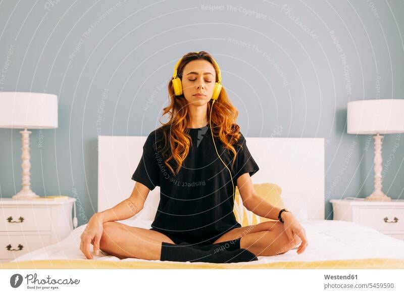 Young woman meditating on bed at home with headphones human human being human beings humans person persons celibate celibates singles solitary people