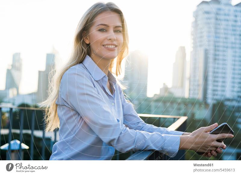 Blonde smiling business woman leaning onto handrail holding smartphone on city rooftop businesswoman businesswomen business women rested on house top Roof Top