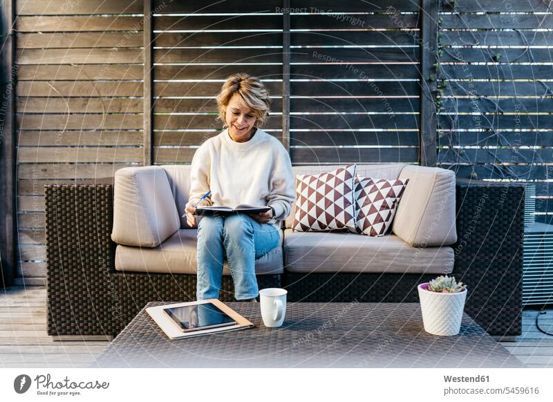 Smiling woman writing in diary while sitting on sofa at rooftop garden color image colour image outdoors location shots outdoor shot outdoor shots day