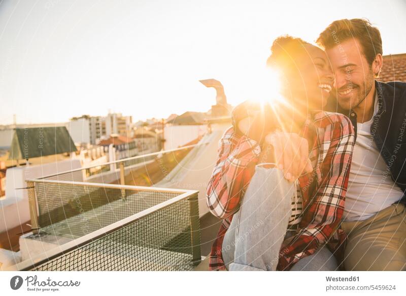 Happy affectionate young couple sitting on rooftop at sunset relax relaxing celebrate partying cuddle snuggle snuggling smile Seated embrace Embracement hug