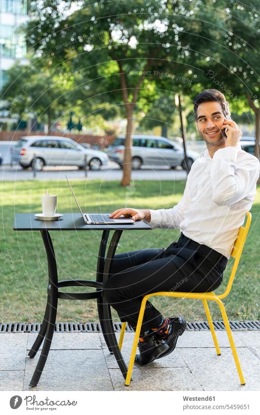 Handsome businessman with laptop on table talking over mobile phone at outdoor cafe color image colour image Spain outdoors location shots outdoor shot