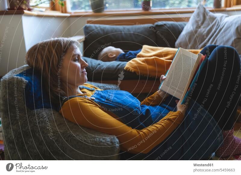 Woman reading book in living room with little son sleeping books couches settee settees sofa sofas chairs Arm Chair Arm Chairs armchairs relax relaxing asleep