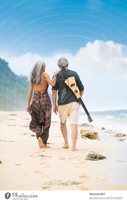 Back view of senior hippie couple with guitar strolling side by side on the beach Hippy hippies flower children Flower power beaches paralell Juxtaposed