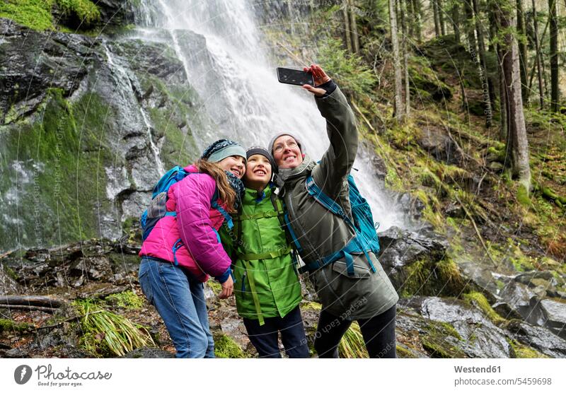 Smiling woman taking selfie with children against Zweribach waterfall in Northern Black Forest color image colour image Germany outdoors location shots