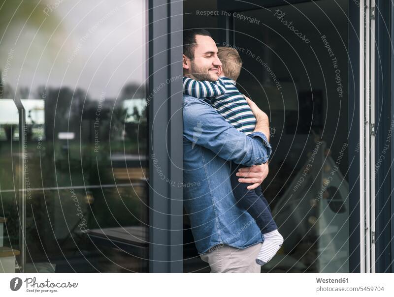 Father hugging son at terrace door at home patio door French window cuddling father fathers daddy dads papa carrying sons manchild manchildren parents family