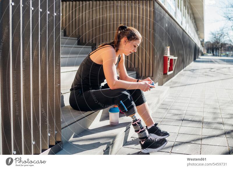 Sporty young woman with leg prosthesis sitting on stairs in the city using smartphone telecommunication phones telephone telephones cell phone cell phones