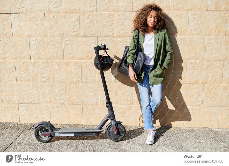 Confident young woman standing with electric push scooter on sidewalk against wall color image colour image outdoors location shots outdoor shot outdoor shots