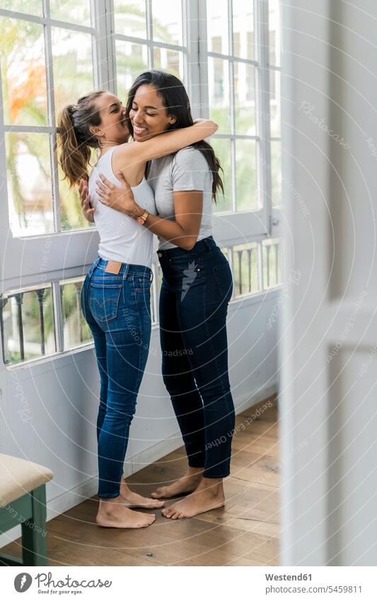 Two happy girlfriends hugging at the window at home female friends windows woman females women happiness embracing embrace Embracement mate friendship Adults