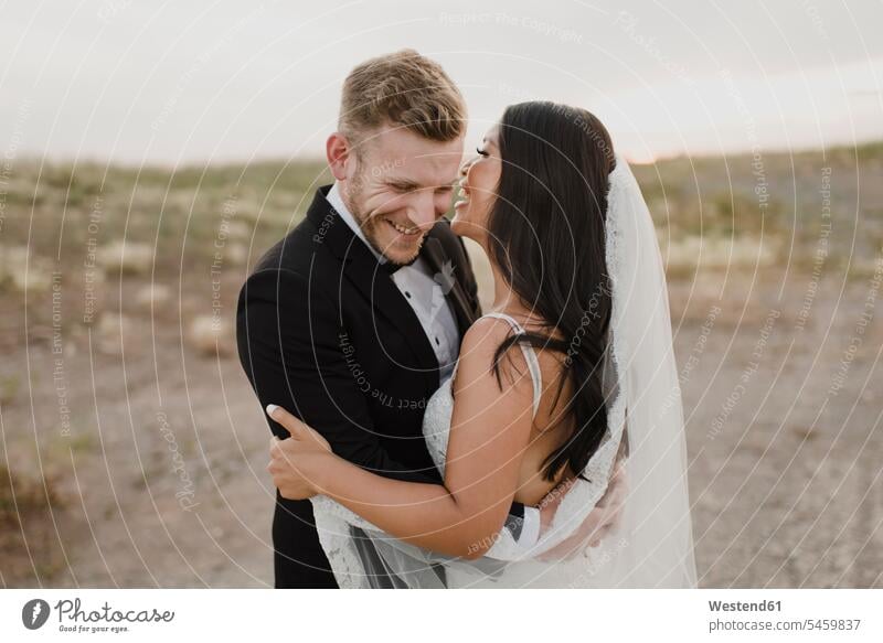 Happy groom embracing bride while standing in field against sky color image colour image outdoors location shots outdoor shot outdoor shots dusk