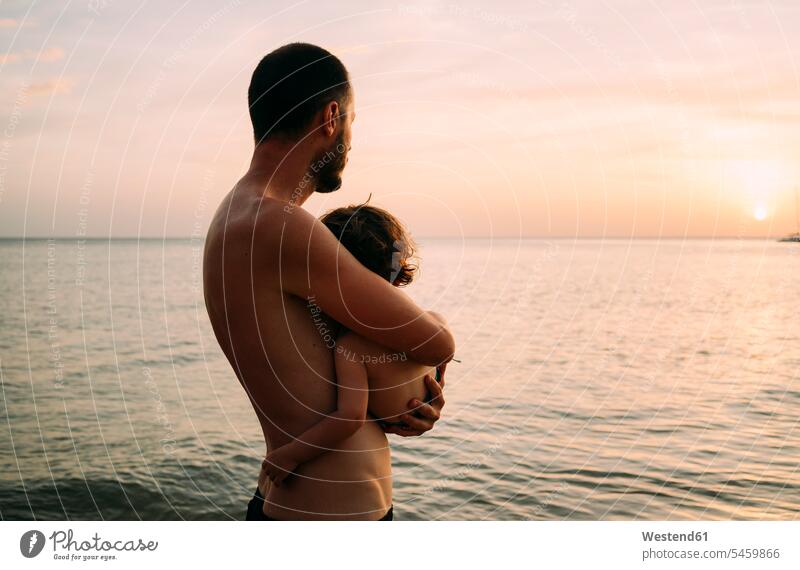 Father holding little daughter on his arms while watching sunset together at the sea sunsets sundown looking looking at father pa fathers daddy dads papa