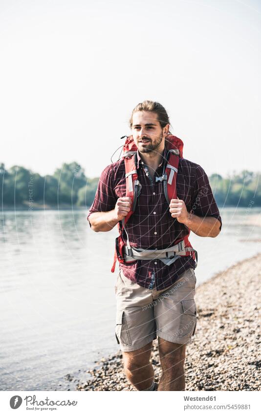 Young man with backpack walking at the riverside rucksacks backpacks back-packs River Rivers going men males riverbank water waters body of water Adults