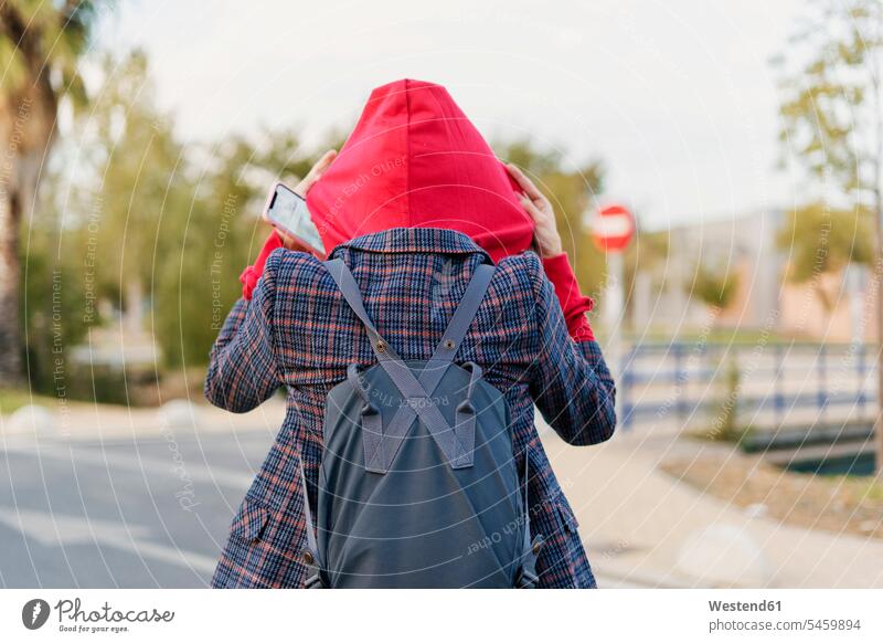 Back view of young woman with backpack and mobile phone putting on hood human human being human beings humans person persons caucasian appearance