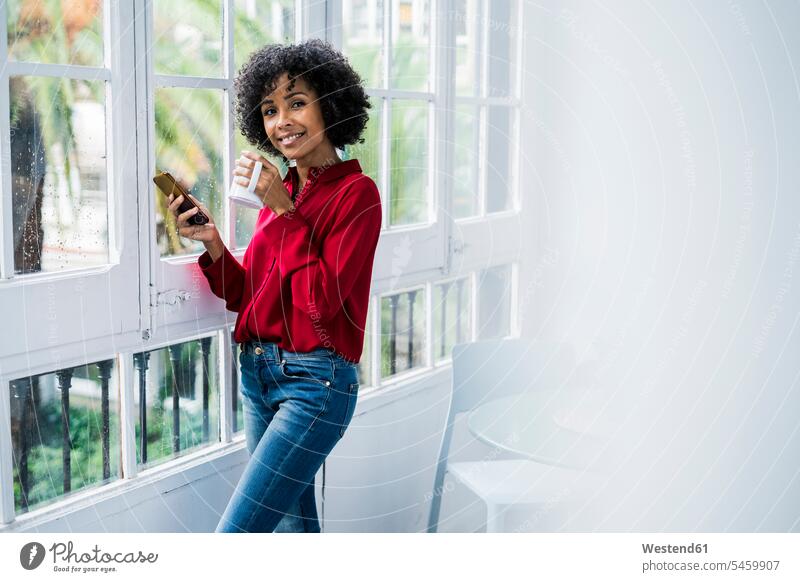 Portrait of smiling woman with cup of coffee and cell phone standing at the window at home windows Coffee Cup Coffee Cups smile females women portrait portraits