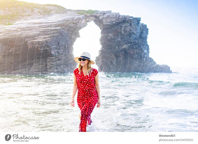 Blond woman wearing red dress and hat and walking along beach, Natural Arch at Playa de Las Catedrales, Spain Eye Glasses Eyeglasses specs spectacles