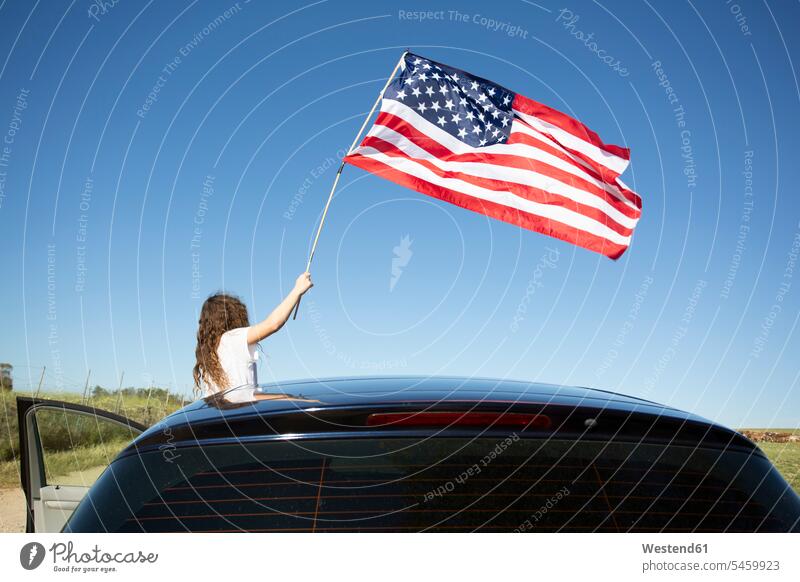 Girl holding American flag out of a car under blue sky skies american flags banner banners automobile Auto cars motorcars Automobiles girl females girls colour