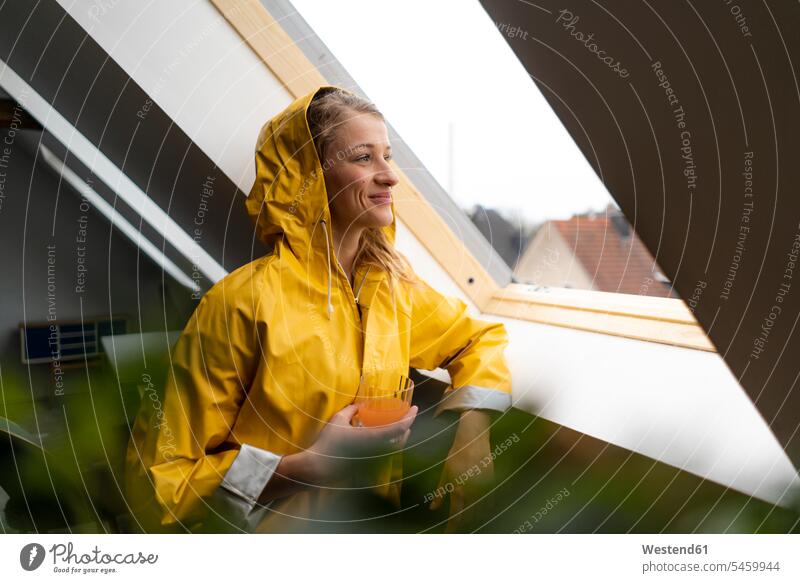 Smiling young woman in raincoat looking out of attic window single singles celibate solitary people celibates solitary person rain coat rain jacket rain slicker