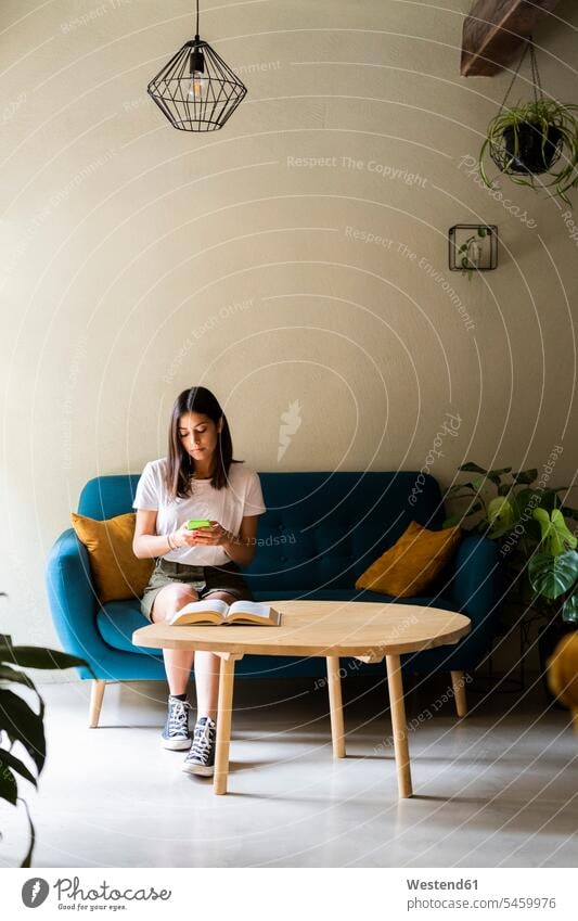 Young woman sitting on a couch using cell phone books T- Shirt t-shirts tee-shirt couches settee settees sofa sofas telecommunication phones telephone