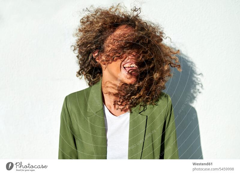 Playful woman tossing curly hair against white wall during sunny day color image colour image outdoors location shots outdoor shot outdoor shots daylight shot