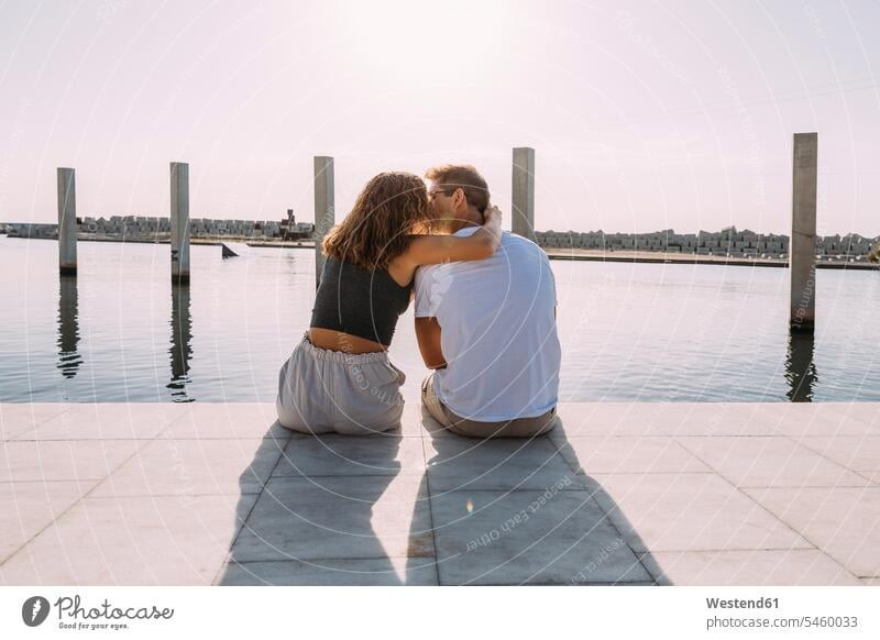 Rear view of young couple sitting on a pier at the sea kissing touristic tourists kisses Seated embrace Embracement hug hugging seasons summer time summertime