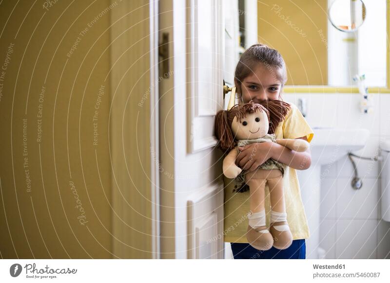 Portrait of smiling little girl standing in doorframe at home holding a doll females girls portrait portraits dolls toilet toilets restrooms child children kid