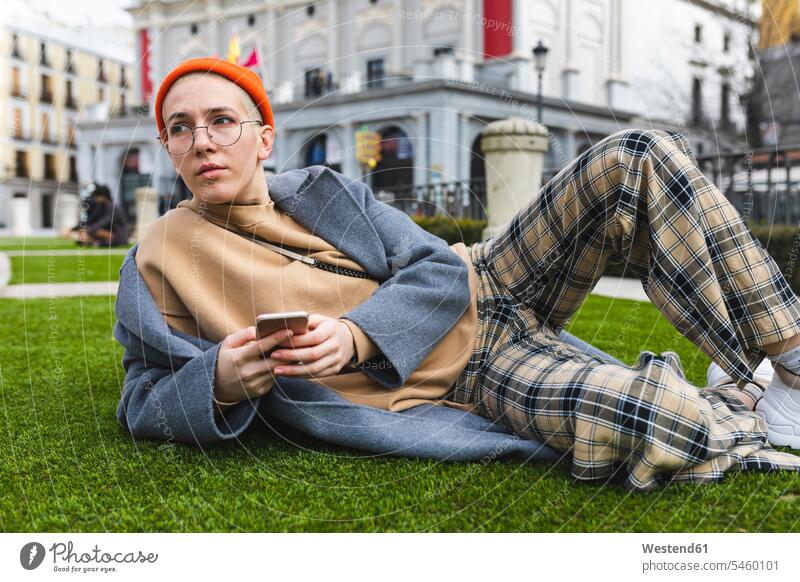 Young woman lying on meadow, holding her phone and looking away Hipster Hipsters Smartphone iPhone Smartphones cap hats caps laying down lie lying down females