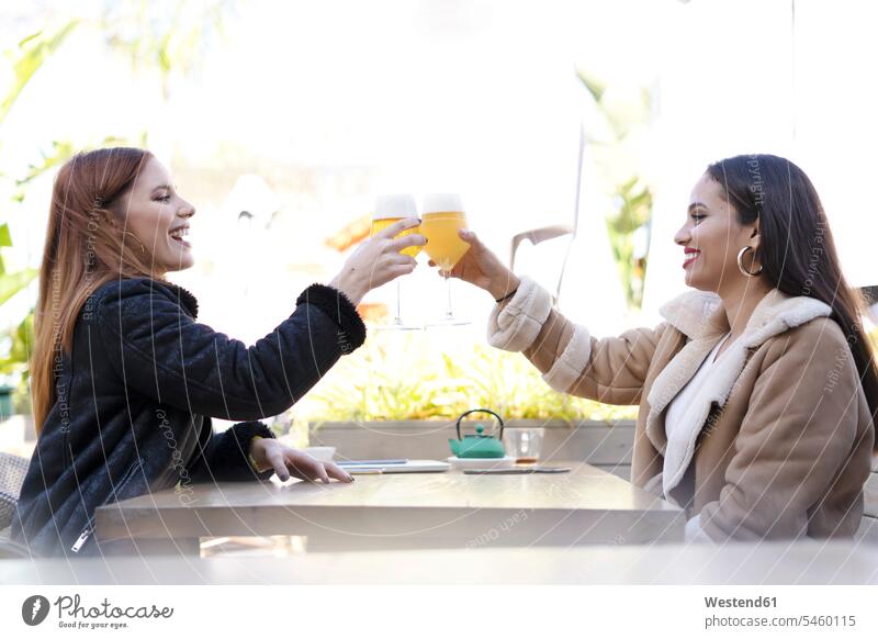 Two female friends clinking beer glasses outdoors at a cafe mate Drinking Glass Drinking Glasses Beer Glasses coat coats jackets Tables hold smile Seated sit
